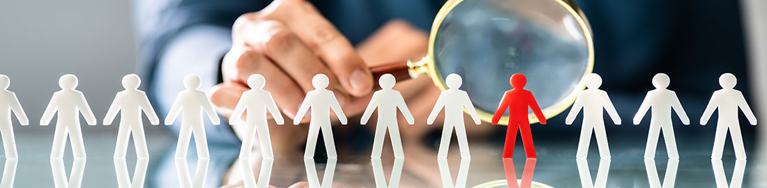 row of white human forms  with one red one on table and person examining red human form with magnifying glass