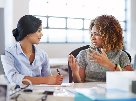 two professional women at desk having conversation, one gesturing with hands