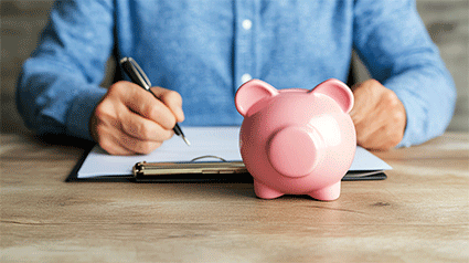 pink piggy bank on desk; man sitting behind desk writing on document with pen